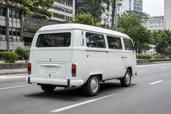 Kombi branca em deslocamento numa avenida de São Paulo, vista de três quartos traseira